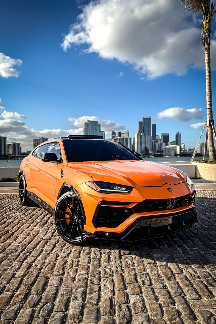 Orange Lamborghini Urus parked on cobblestones with a waterfront city skyline behind it